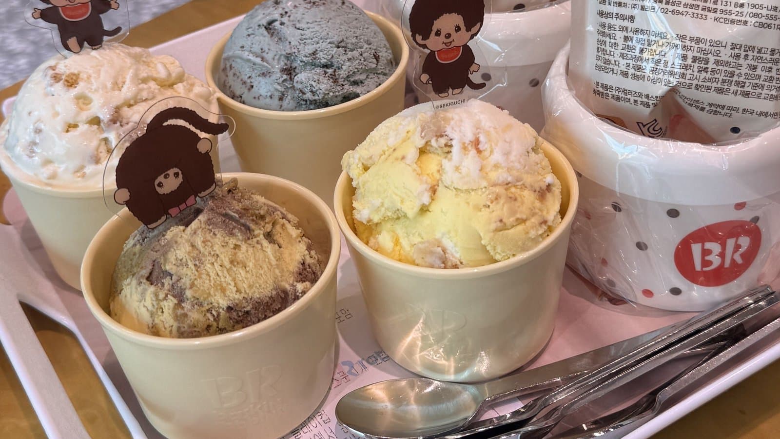 Four Baskin-Robbins ice cream cups topped with Monchhichi character deco picks on a pink tray in a Korean store
