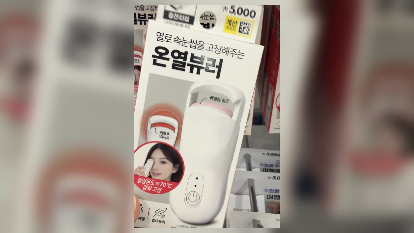 Daiso's 5,000 won heated eyelash curler box held in hand at a Daiso store shelf, showing the white curler with pink heat-indicator pad and 70C curling temperature